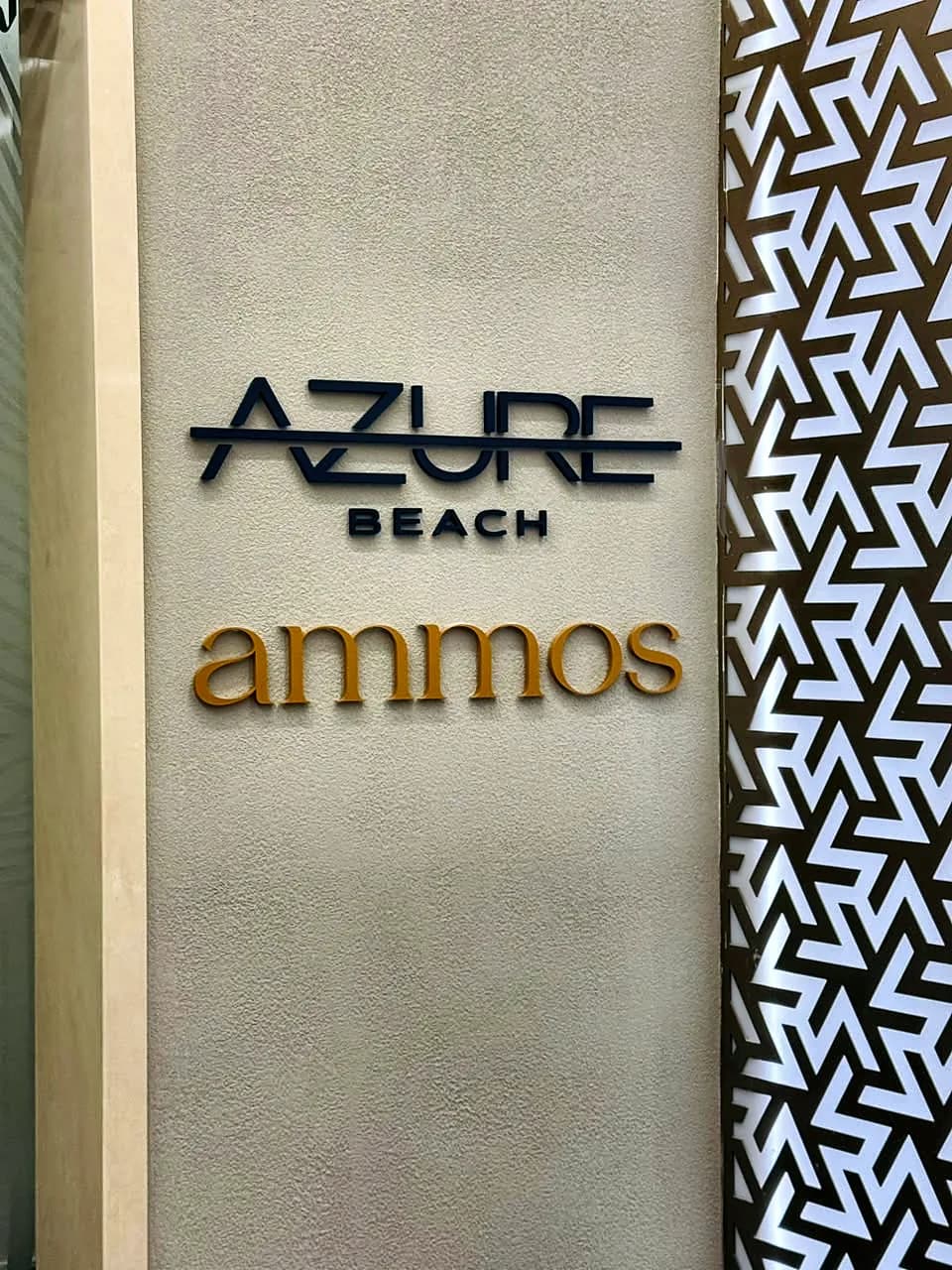 Raised dark Azure Beach lettering above gold ammos lettering on a pale wall beside a patterned screen