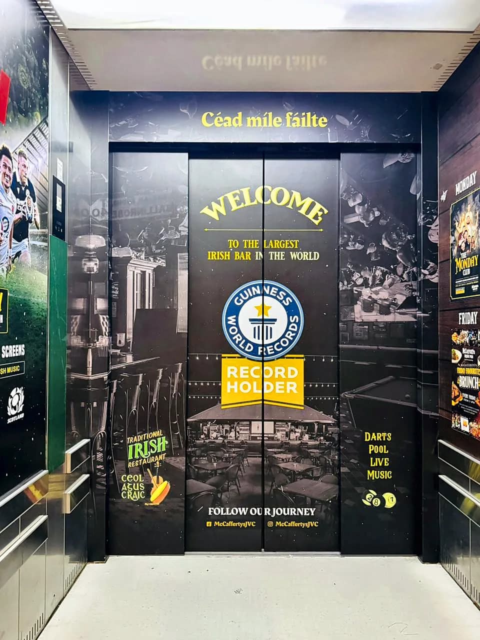 Lift lobby wrapped in dark McCafferty's graphics with Céad míle fáilte above the doors, a Welcome to the largest Irish bar in the world message and a Guinness World Records record holder badge