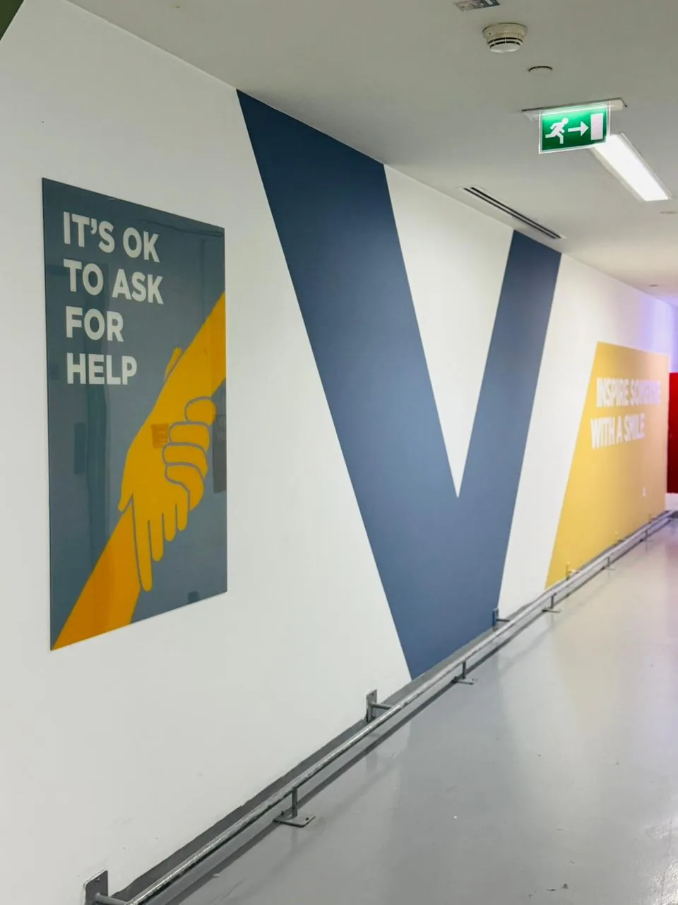 Staff corridor wall with a large blue V graphic, a yellow Inspire someone with a smile panel and a printed acrylic sign reading It's OK to ask for help