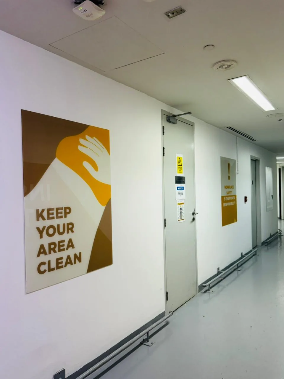 Printed acrylic sign reading Keep your area clean and a workplace safety panel along a staff corridor