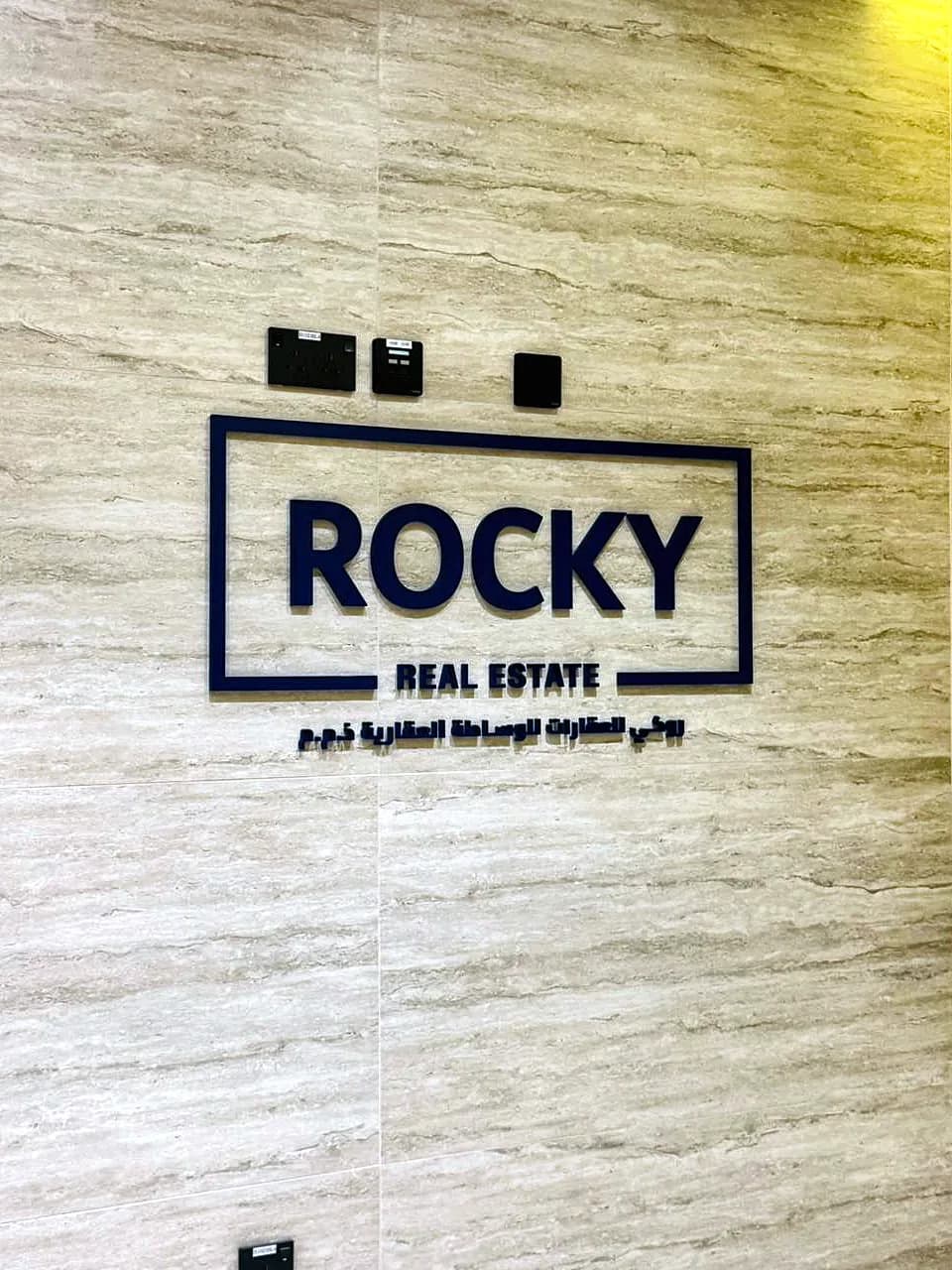 Raised navy Rocky Real Estate logo sign with Arabic text below, fixed to a stone wall