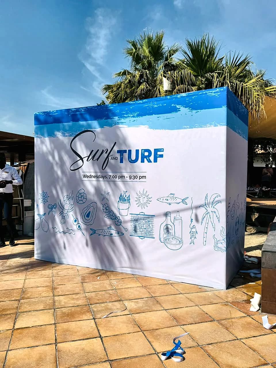 Outdoor booth wrapped in white and blue Surf and Turf graphics with hand drawn seafood illustrations and the time Wednesdays 7:00 pm to 9:30 pm